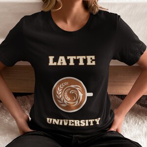 Può includere: Maglietta nera con il testo "LATTE UNIVERSITY" in lettere dorate. Un'immagine stilizzata di una tazza di caffè con latte art è centrata sulla maglietta.