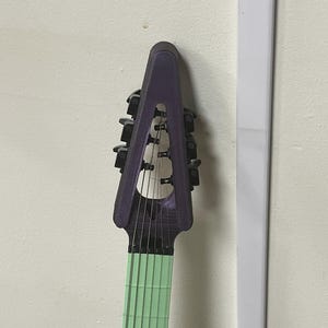May include: A unique, purple electric guitar headstock with black tuning pegs. The fretboard is a light green color. The guitar is mounted on a white wall.