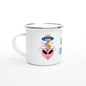 May include: White enamel mug with a quirky design. The mug features a pink heart-shaped alien with black eyes, a crescent moon, and a UFO above it. The UFO emits colourful rays. The text "Stay Bright" is printed on the side.