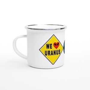 Könnte beinhalten: Weißer Emaille-Becher mit silbernem Rand und Henkel. Der Becher hat eine gelbe Raute mit den Worten "WE URANUS" und einem roten herzförmigen Alien-Gesicht. Eine blau-weiße Grafik ist auf der Seite sichtbar.