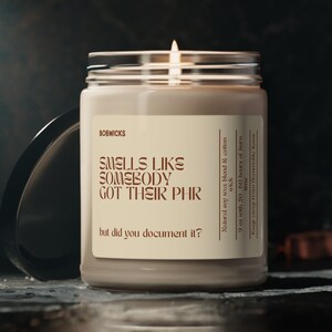 May include: A beige candle with a glass jar and a black lid. The candle is lit and has a brown label with the text "SMELLS LIKE SOMEBODY GOT THEIR PHR but did you document it?"  Natural soy wax blend & cotton wick with 50 hours of burn.