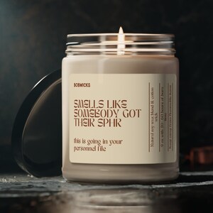 May include: A beige candle with a glass jar and a black lid. The candle is labeled "Smells Like Somebody Got Their Sphr" and "This is going in your personnel file".