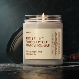 May include: A beige candle jar with a black lid. The candle is labeled "Smells like somebody got their shrm-scp" and "This is going in your personnel file". The candle is made with natural soy wax and cotton wick and has a burn time of 50-60 hours.