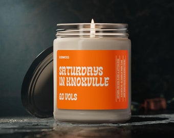 May include: A lit candle in a glass jar with a black lid. The jar has an orange label with the text "Saturdays in Knoxville" and "Go Vols" in white. The candle is on a dark, textured surface.