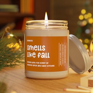 May include: A lit candle in a glass jar with a lid. The jar has an orange label that reads "Smells like Fall" and "infused with the scent of Pumpkin Spice and basic bitches". The candle is on a wooden surface.