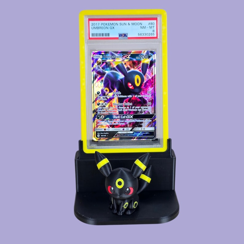 Puede incluir: Una figura negra de Umbreon con detalles amarillos se encuentra frente a una carta de Pok&eacute;mon calificada. La carta, en una funda amarilla, presenta a Umbreon GX y es del set Pok&eacute;mon Sol y Luna de 2017.