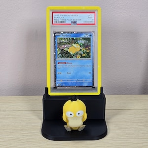 May include: A yellow and black display stand holding a graded Pok&eacute;mon card featuring Psyduck. The card is encased in a yellow holder with text that reads "2025 POKEMON MEP EN PSYDUCK." A small Psyduck figurine sits in front of the card.