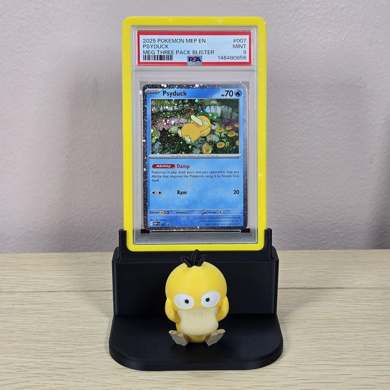 May include: A yellow and black display stand holding a graded Pok&eacute;mon card featuring Psyduck. The card is encased in a yellow holder with text that reads "2025 POKEMON MEP EN PSYDUCK." A small Psyduck figurine sits in front of the card.