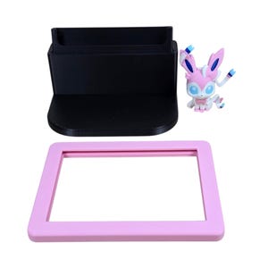 May include: A black phone stand, a pink rectangular frame, and a Sylveon figurine are displayed against a white background. The Sylveon figure is pink, white, and blue. The phone stand has a slot for a phone.