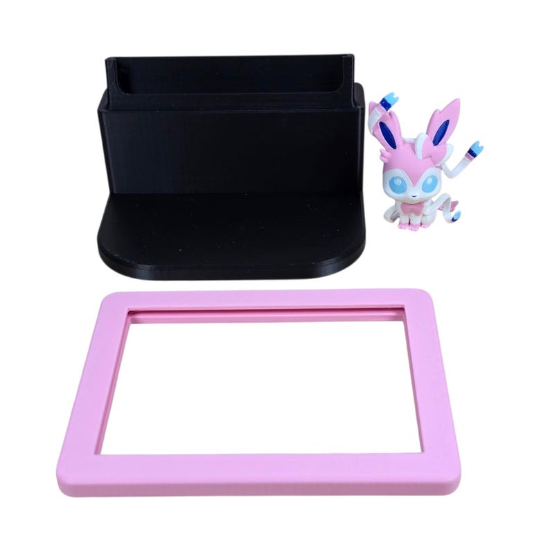 May include: A black phone stand, a pink rectangular frame, and a Sylveon figurine are displayed against a white background. The Sylveon figure is pink, white, and blue. The phone stand has a slot for a phone.