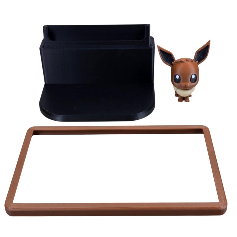 May include: A black rectangular stand with a slot, a brown rectangular frame, and a small brown Eevee figurine with large ears and a white collar. The items are on a white background.