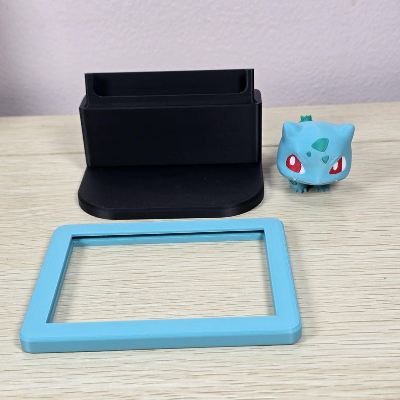 Puede incluir: Un soporte negro impreso en 3D con una base rectangular y una ranura, un marco rectangular azul claro y una peque&ntilde;a figura de Bulbasaur. El Bulbasaur es azul claro con ojos rojos y un bulbo verde en la espalda.