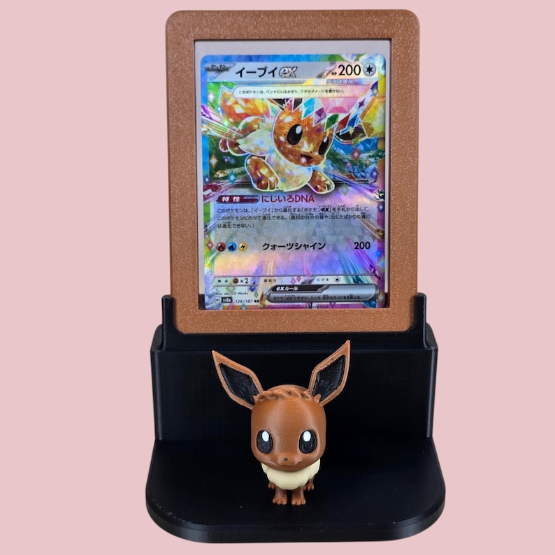 May include: A brown and black display stand holding a Pok&eacute;mon trading card featuring a colorful Eevee character. A small Eevee figurine is positioned below the card. The stand is brown and black.
