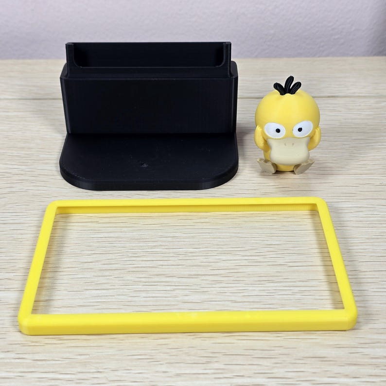 May include: A black phone stand, a yellow rectangular frame, and a yellow Psyduck figurine are arranged on a light wood surface. The phone stand has a rectangular base and a slot for holding a phone. The Psyduck figurine is yellow with a white beak and large eyes.