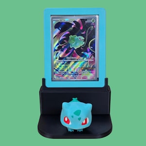 Puede incluir: Una figura de Bulbasaur turquesa se encuentra debajo de una tarjeta Pok&eacute;mon enmarcada en turquesa. La tarjeta presenta una ilustraci&oacute;n de Bulbasaur con el texto "Bulbasaur" y "Bind Down". La exhibici&oacute;n est&aacute; sobre un fondo verde.