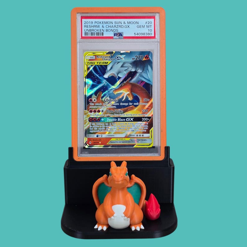 May include: A graded Pok&eacute;mon card in an orange case, featuring Reshiram & Charizard GX from the 2019 Sun & Moon Unbroken Bonds set. A small orange Charizard figure with green wings and a red flame sits below the card.