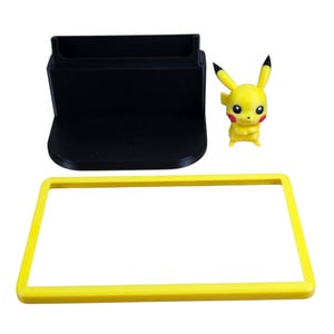 May include: A black plastic stand with a rectangular base and a slot, a yellow rectangular frame, and a yellow Pikachu figurine. The Pikachu figure has red cheeks and black-tipped ears. The items are on a white background.