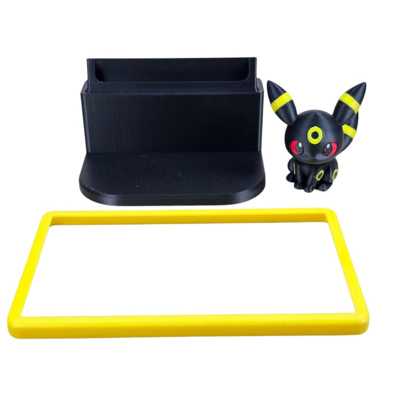 Puede incluir: Un organizador de escritorio negro con un marco rectangular amarillo y una figura de Umbreon negra. La figura de Umbreon es negra con detalles amarillos y ojos rojos. El organizador tiene una ranura para guardar objetos.