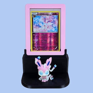 May include: A pink framed Pok&eacute;mon card featuring Sylveon, displayed on a black stand with a Sylveon figurine. The card has a colorful design with the name "Sylveon" at the top. The figurine is pink, white, and blue.