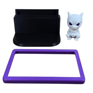 May include: A black phone stand, a purple rectangular frame, and a white figurine of a creature with purple eyes are displayed against a white background. The phone stand has a slot for a phone. The figurine is in a squatting position.