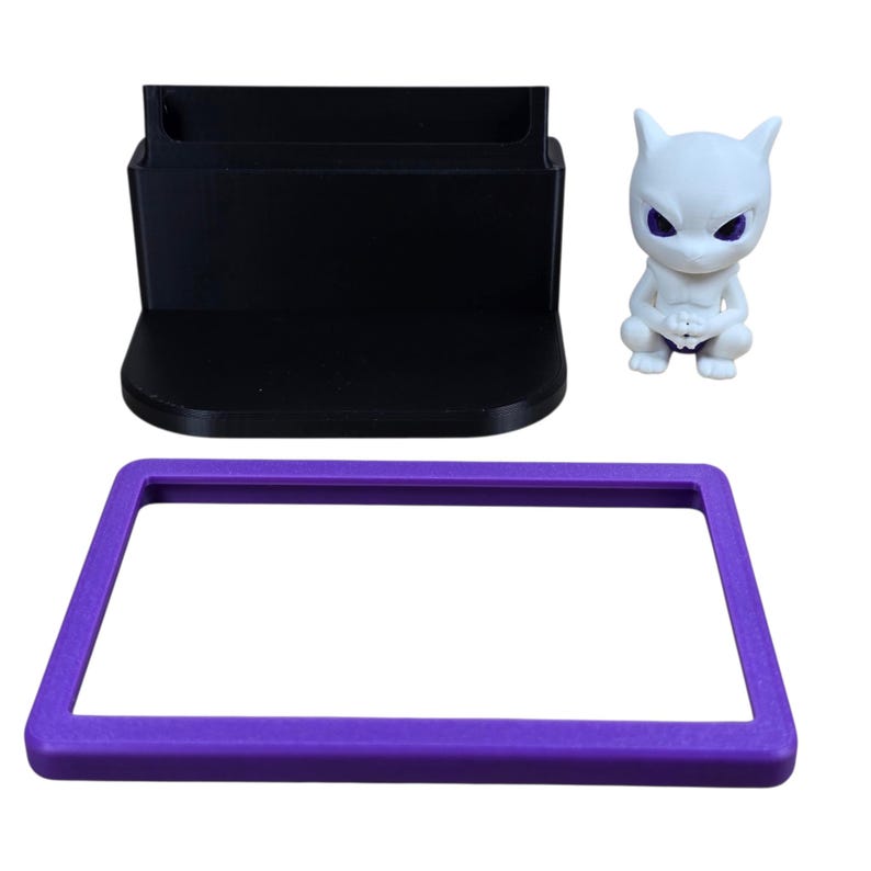 May include: A black phone stand, a purple rectangular frame, and a white figurine of a creature with purple eyes are displayed against a white background. The phone stand has a slot for a phone. The figurine is in a squatting position.
