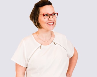 Puede incluir: Una camisa blanca de manga corta con cuello redondo y dos cremalleras plateadas en los lados. La camisa está hecha de una tela ligera y tiene un corte holgado.