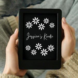 Könnte beinhalten: Ein schwarzer E-Reader mit einem weißen Gänseblümchenmuster und dem Text "Jessica's Kindle" auf dem Bildschirm.