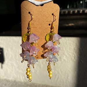 K&ouml;nnte beinhalten: Ohrringe mit Anh&auml;ngern mit Clustern aus durchscheinenden lila und blauen Blumenanh&auml;ngern mit goldfarbenen Akzenten. Die Ohrringe haben auch kleine gelbe Perlenanh&auml;nger und gr&uuml;ne Blattdetails.