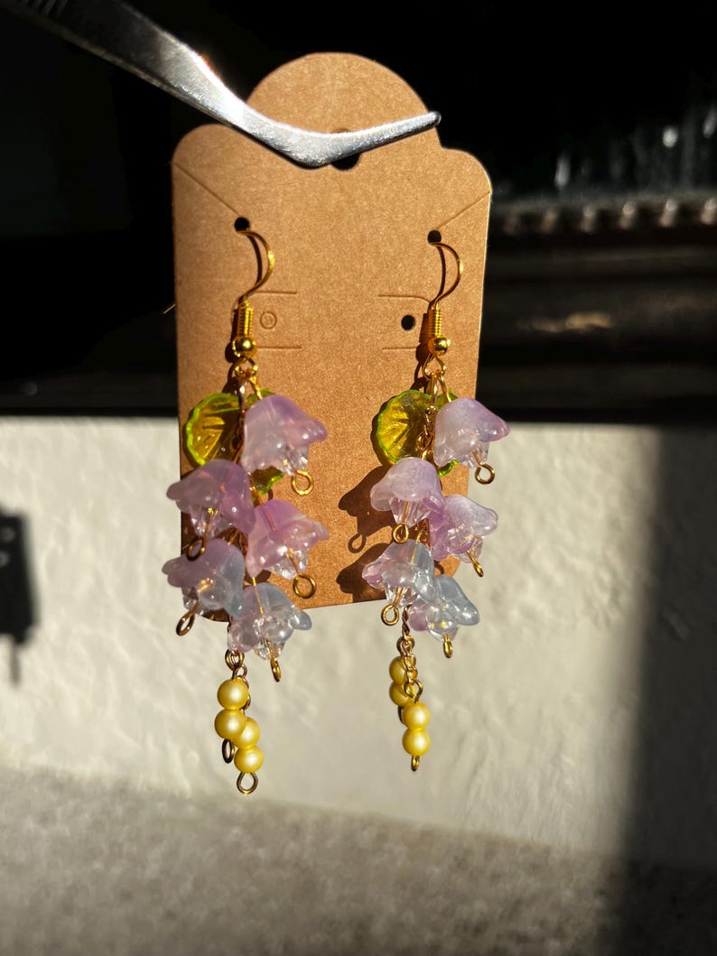 K&ouml;nnte beinhalten: Ohrringe mit Anh&auml;ngern mit Clustern aus durchscheinenden lila und blauen Blumenanh&auml;ngern mit goldfarbenen Akzenten. Die Ohrringe haben auch kleine gelbe Perlenanh&auml;nger und gr&uuml;ne Blattdetails.