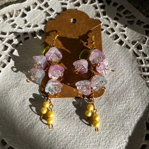 K&ouml;nnte beinhalten: H&auml;ngeohrringe mit Clustern aus durchscheinenden lila und wei&szlig;en Blumenperlen, goldfarbenen Dr&auml;hten und gelben Perlenclustern. Die Ohrringe werden auf einem braunen Etikett pr&auml;sentiert. Ein zartes Blumendesign.