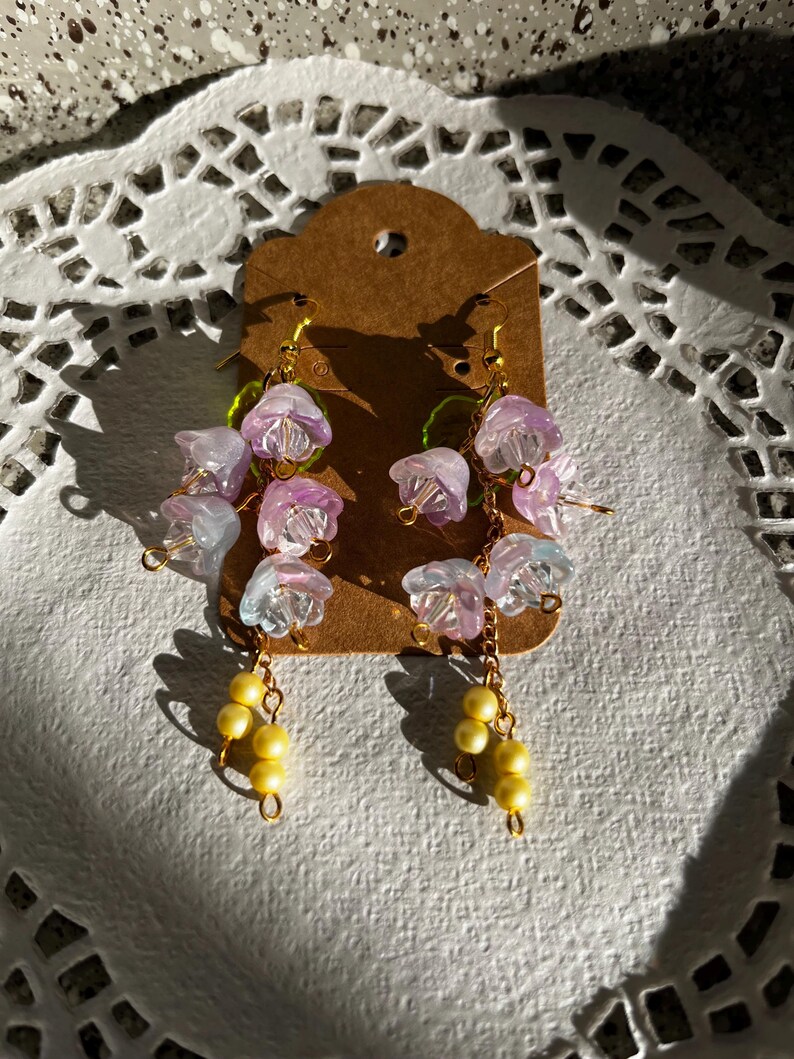 K&ouml;nnte beinhalten: H&auml;ngeohrringe mit Clustern aus durchscheinenden lila und wei&szlig;en Blumenperlen, goldfarbenen Dr&auml;hten und gelben Perlenclustern. Die Ohrringe werden auf einem braunen Etikett pr&auml;sentiert. Ein zartes Blumendesign.