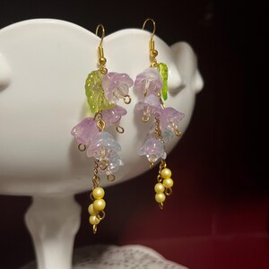K&ouml;nnte beinhalten: Ohrringe zum Aufh&auml;ngen mit durchscheinenden, glockenf&ouml;rmigen Bl&uuml;ten in Lila und Blau, goldfarbenen Haken, kleinen gelben Perlenclustern und gr&uuml;nen Blattakzenten. Ein charmantes Blumendesign.