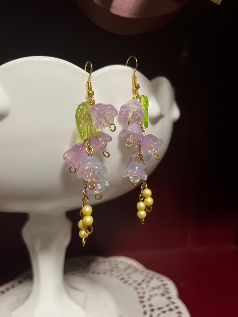 K&ouml;nnte beinhalten: Ohrringe zum Aufh&auml;ngen mit durchscheinenden, glockenf&ouml;rmigen Bl&uuml;ten in Lila und Blau, goldfarbenen Haken, kleinen gelben Perlenclustern und gr&uuml;nen Blattakzenten. Ein charmantes Blumendesign.