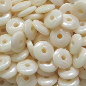 May include: A collection of off-white, donut-shaped beads with a pearlescent sheen. The beads have a hole in the center and are scattered across the image. A strand of similar beads is shown at the bottom.