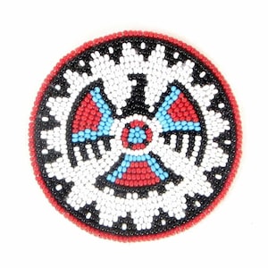 May include: A round beaded design with a central motif of four red, blue, and white triangles. The design is surrounded by a black and white beaded border with a red outer edge.