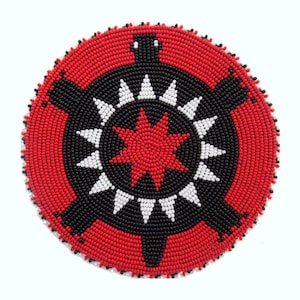 May include: A circular beaded design with a turtle motif. The design uses red, black, and white beads. The outer ring is red, with a black turtle shape in the centre, and a red starburst in the middle. It is likely a decorative item.