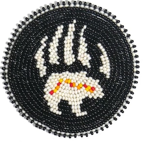 May include: A round, beaded coaster with a black background and a white bear paw print. The bear paw print is made of white beads, with yellow and red accents. The border is black with a white beaded edge.