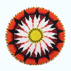 May include: Circular beaded design with a floral pattern. The center is yellow, surrounded by white petals, and a red, black, and orange border. The design is likely a coaster or decorative piece.