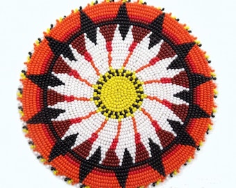 May include: Circular beaded design with a floral pattern. The center is yellow, surrounded by white petals, and a red, black, and orange border. The design is likely a coaster or decorative piece.