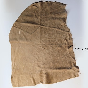 May include: A piece of tan-colored leather, approximately 17 inches by 12.5 inches, with a rough, uneven edge. The leather has a textured surface and appears to be pliable. The image shows the leather against a white background.