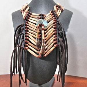 May include: A handcrafted necklace with a Native American design. The necklace features light brown wooden beads, dark brown leather, and copper-colored accents. A central pendant with a blue-green stone and leather fringe adds detail. The necklace is displayed on a black mannequin.