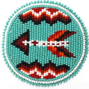 May include: A round beaded design with turquoise, red, white, and black beads. The design features two stylized birds with wings spread.