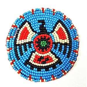 May include: A round beaded design with a blue background, black, white, red, and green beads. The design features a central circle with four radiating arms.