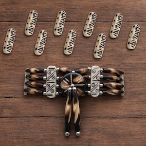 May include: A handcrafted choker necklace with a central circular design and dangling accents. The necklace features a combination of brown and cream-colored beads and decorative elements. Ten cream-colored, oblong beads with black geometric patterns are scattered above the necklace.