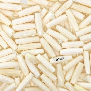 May include: Ivory-colored barrel beads, approximately 1 inch long, are scattered across the frame. The beads have a smooth, glossy finish and are cylindrical with slightly rounded ends. The image shows a large quantity of these beads.