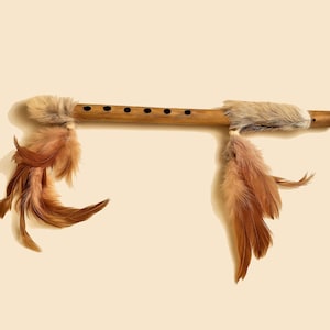 May include: A wooden flute with six finger holes and decorative accents. The flute has a light brown color with fur and feathers attached at each end. The feathers are shades of brown and tan.