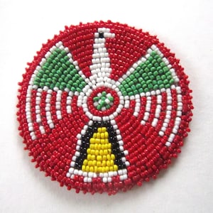 May include: A round beaded design with a red, white, green, yellow, and black color scheme. The design features a central bell shape with a white and black outline.