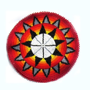May include: A round, beaded decorative item with a vibrant sunburst design. The central circle is white, bisected by black lines. Black, yellow, and gray triangles radiate from the center, set against a red and orange background. The outer edge is a solid red.