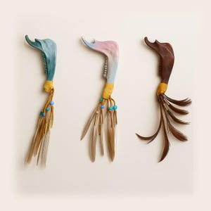 May include: Three decorative jawbone ornaments. One is blue, one is pink and blue, and one is brown. Each has a yellow band, beads, and feathers or plumes. The ornaments are displayed against a white background.