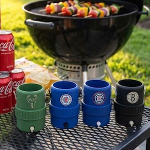 May include: A backyard barbecue scene with a black grill, skewers of grilled vegetables, and a stack of Coca-Cola cans. Several beverage coolers with team logos are on a metal table, ready for a summer gathering.
